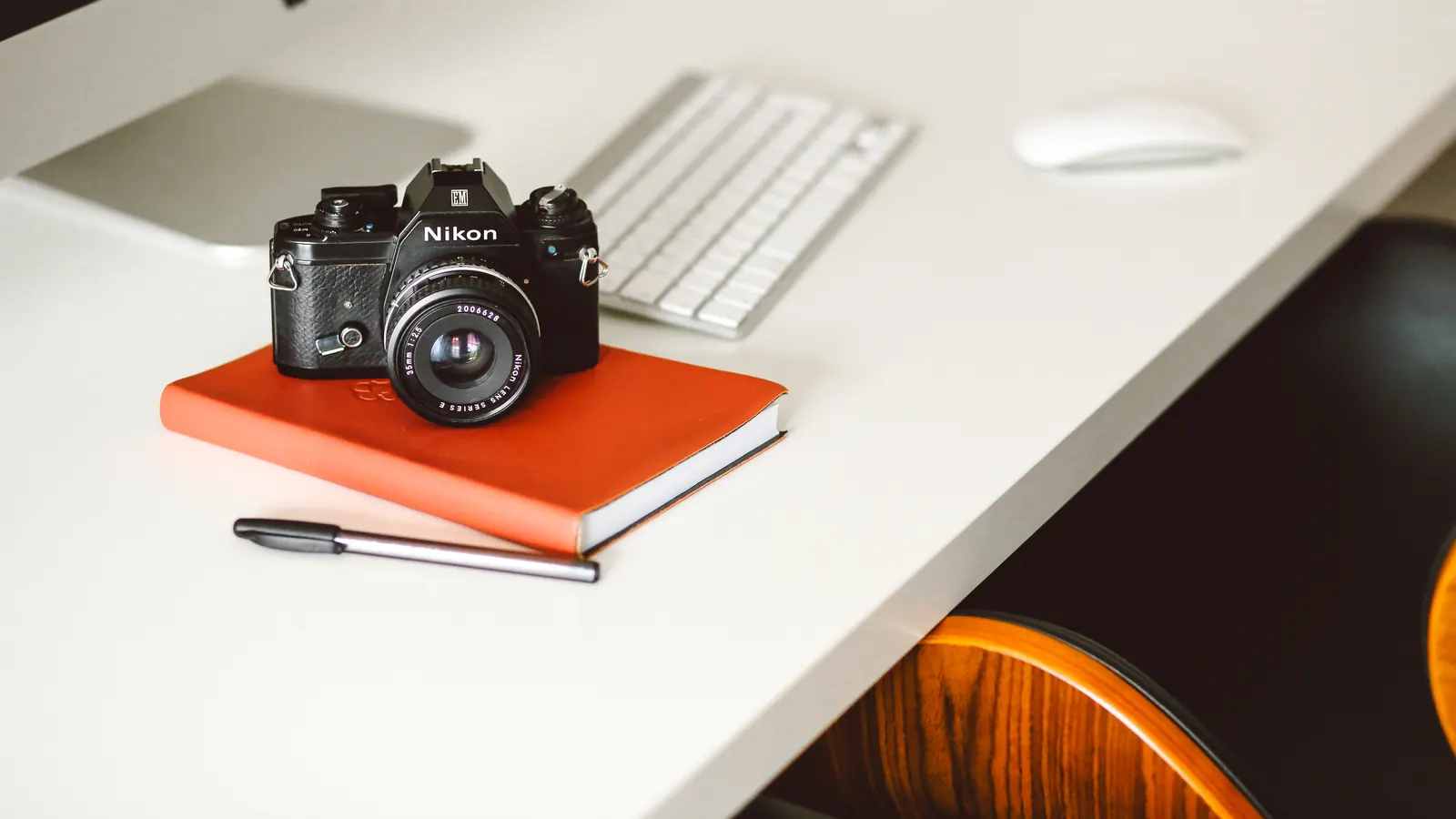 A black film camera resting on an orange notebook on a white desk, beside a pen, a keyboard and a mouse