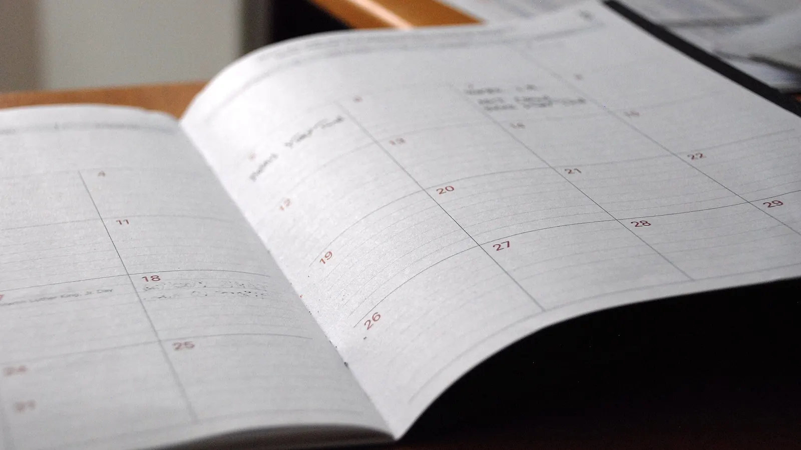 An open paper monthly planner on a wooden desk, its date squares mostly empty