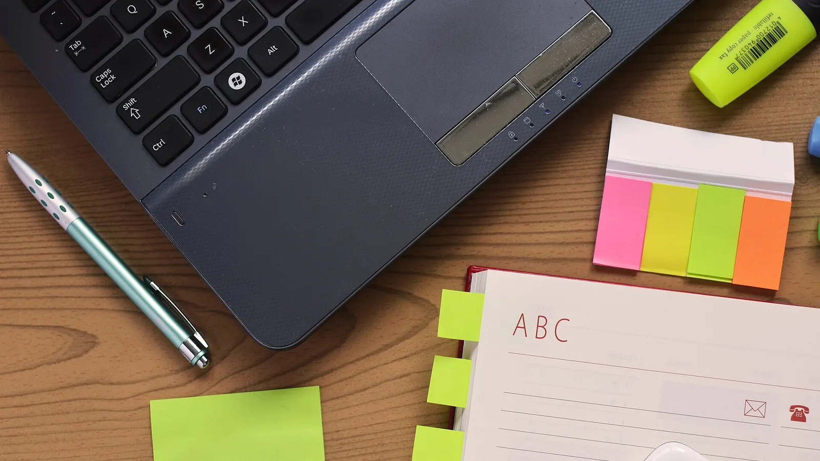 A laptop on a wooden desk beside a pen, a notebook with coloured tabs and a stack of pink, yellow, green and orange sticky tabs