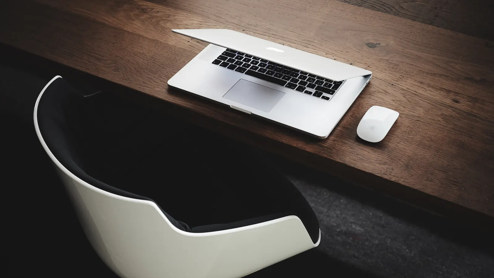 A laptop and a mouse on a dark wooden desk, with an empty white chair pulled up to it